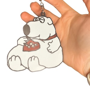 Peut inclure: Porte-clés représentant un chien de dessin animé blanc avec un motif en forme de cœur rouge et blanc sur la poitrine. Le chien a un nez gris, des lunettes et un collier rouge. Fond blanc.