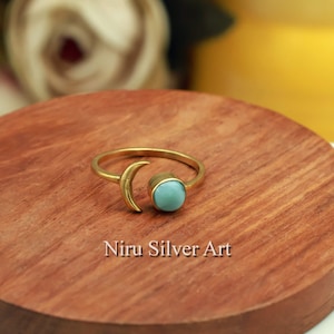 Puede incluir: Un anillo dorado con un diseño de media luna y una piedra preciosa redonda de color azul claro. El anillo está sobre una superficie de madera, con el texto "Niru Silver Art" visible debajo.