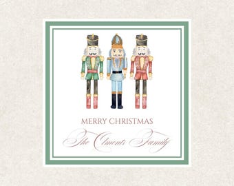 May include: A square Christmas card with three watercolour nutcracker soldiers in green, blue, and red uniforms. The text "MERRY CHRISTMAS" and a family name are displayed in elegant script within a green border.