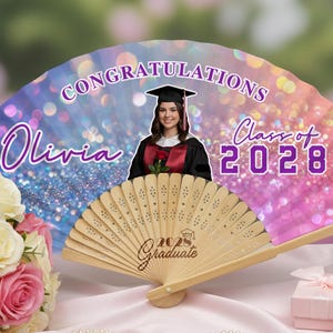 Puede incluir: Un abanico de graduación con el texto "Felicidades Olivia Clase de 2028" en morado y blanco. El abanico presenta una foto de una graduada con birrete y toga. Un abanico de madera está en primer plano con el texto "2028 Graduado".