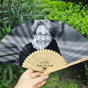 Op de afbeelding: Een gepersonaliseerde herdenkingsventilator met een zwart-witfoto van een vrouw met een bril. De ventilator heeft een houten handvat en bevat de tekst "IN LOVING MEMORY Emily R. Smith 1948-2026" en "YOUR CUSTOM TEXT".