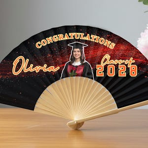 May include: A personalized graduation fan with the name "Olivia" in gold script. The fan features a photo of a graduate in a cap and gown, with the words "Congratulations" and "Class of 2028" in orange. The fan is black with a wooden handle.