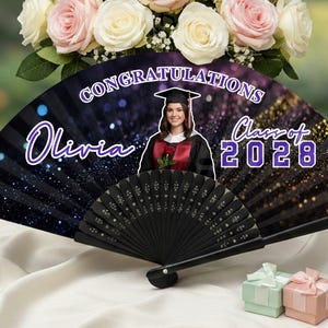 May include: A black graduation fan with the text "Congratulations Olivia Class of 2028" and a photo of a graduate. The fan is decorated with glitter and is set against a backdrop of flowers and gifts.