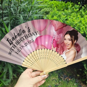 May include: A personalised hand fan featuring a woman and pink balloons. The fan is light pink with the text "Isabella's 30TH BIRTHDAY 12.18.2025". It has a wooden handle and is held open.