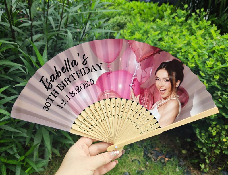 May include: A personalised hand fan featuring a woman and pink balloons. The fan is light pink with the text "Isabella's 30TH BIRTHDAY 12.18.2025". It has a wooden handle and is held open.