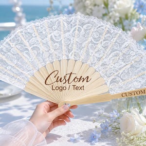 Peut inclure: Un éventail pliant en dentelle blanche avec des baleines en bois clair. L'éventail présente le texte Custom Logo / Text imprimé en brun foncé sur la partie centrale en bois et les mots CUSTOM TEXT sur la baleine latérale de la poignée.