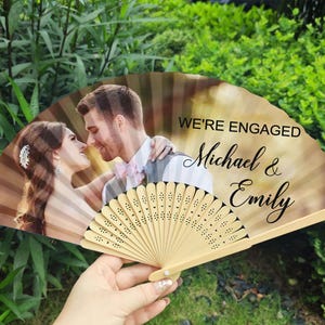 May include: A beige folding fan with a photo of a couple, with the text "WE'RE ENGAGED Michael & Emily". The fan is held in front of a green leafy background. The fan is made of wood and paper.