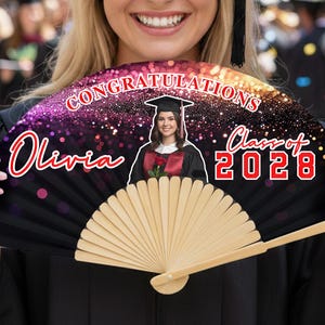 Op de afbeelding: Een zwarte afstudeerwaaier met een foto van een afgestudeerde, de naam "Olivia" en "Class of 2028" in het rood. De waaier heeft een houten handvat en een decoratieve bovenkant met het woord "CONGRATULATIONS" in het wit.