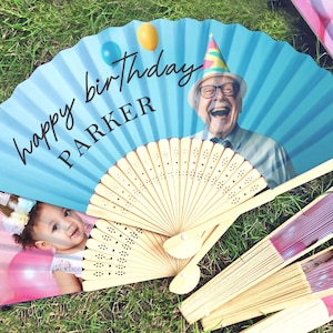 May include: A selection of personalised birthday fans. One fan displays a photo of a laughing senior with a party hat, the text "happy birthday PARKER", and balloons. Another fan shows a baby. Other fans have pink and purple stripes.