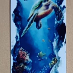 May include: A white bookmark featuring an underwater scene with a sea turtle, fish, and coral. The design is framed by a dark blue border, and the top has a hole for a tassel.