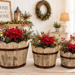 May include: Three rustic wooden buckets with a distressed white finish and dark metal bands, each filled with artificial holiday greenery, red poinsettia flowers, pinecones, and red berries, arranged in a descending size order.
