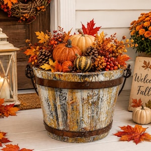 May include: A rustic wooden bucket with a distressed white and brown finish, featuring dark metal bands and handles. The bucket is filled with an arrangement of orange pumpkins, gourds, pinecones, red berries, and vibrant autumn maple leaves.