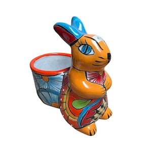 May include: A ceramic rabbit planter with orange, blue, red, and green accents. The rabbit has blue ears and eyes, and the planter is decorated with floral patterns. The planter is designed to hold small plants or flowers.