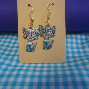 May include: A pair of dangle earrings featuring cat-shaped charms with gold-tone metal hooks. The cat charms are decorated with a colorful geometric pattern in shades of blue, pink, green, and white, with a distinct segmented design on the body.