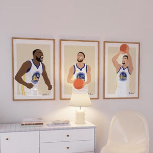 Peut inclure: Trois impressions d'art encadrées représentant des joueurs de basket-ball des Golden State Warriors. Chaque impression a un fond beige clair et un cadre en bois. Les joueurs portent des uniformes blancs et bleus. Les impressions sont affichées au-dessus d'une commode blanche.