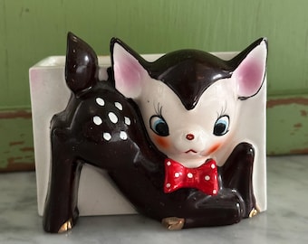 May include: A ceramic planter shaped like a fawn, with a brown body and white spots. The fawn has a white face, blue eyes, and a red polka-dot bow tie. The planter is rectangular and white with gold accents.