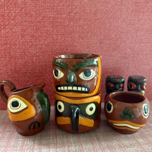 May include: A collection of brown ceramic mugs and salt and pepper shakers with Native American totem pole designs. The mugs feature painted faces with green, orange, and white accents. The salt and pepper shakers have similar designs.