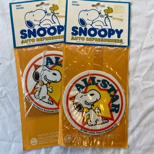 May include: Two packages of car air fresheners featuring Snoopy, the Peanuts character. The air fresheners are round and have a yellow star with the words "All-Star" in red and white. Snoopy is depicted in a red and white baseball uniform with a yellow star on his chest. The packages are yellow and have the words "Snoopy Auto Refreshers" in blue and white.