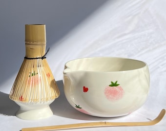 以下が含まれることがあります： 白いセラミックボウル、ピンクのイチゴ模様、竹の泡立て器、竹のスクープを備えた抹茶ティーセット。泡立て器は白いベースで、ピンクのイチゴのデザインで飾られています。