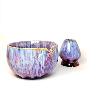 May include: A ceramic bowl and a small vase with a matching glaze. The bowl is round with a spout and a yellow rim. The glaze has a mix of blue, purple, and red colours. The vase is a similar colour and shape.