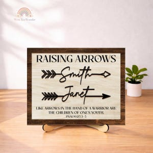 May include: A rectangular wooden sign with a dark brown frame and a light wood-grain center. The text reads RAISING ARROWS, followed by the names Smith and Janet integrated into arrow graphics, and the verse LIKE ARROWS IN THE HAND OF A WARRIOR ARE THE CHILDREN OF ONE'S YOUTH. PSALM 127:3-5.