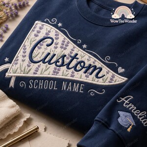 May include: A navy blue sweatshirt featuring an embroidered pennant design with a lavender floral pattern. The pennant displays the text Custom and SCHOOL NAME in dark blue script. The sleeve includes the name Amelia and a graduation cap icon.