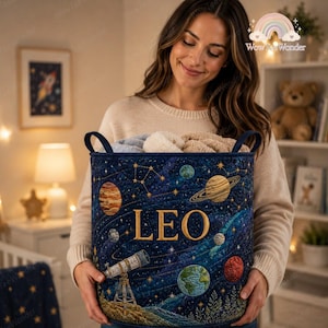 May include: A deep blue quilted fabric storage basket featuring a space-themed print with planets, stars, constellations, and a telescope. The name LEO is embroidered in large gold letters across the center of the basket, which contains soft blankets.
