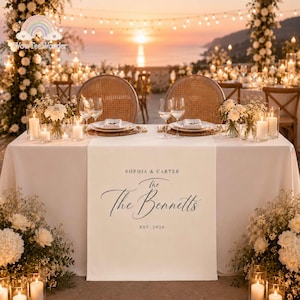 May include: A white fabric table runner featuring black printed text that reads Sophia & Carter, The Bennetts, and Est. 2026. The runner is draped over a table set with white plates, clear wine glasses, and white floral arrangements with candles.