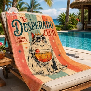 May include: A rectangular beach towel featuring a graphic of a fluffy cat wearing a gold crown and black sunglasses, holding a cocktail with a cherry. The text reads The Desperado Club So Fun It Hurts, set against a vintage-style striped background.