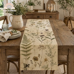 May include: A long rectangular table runner featuring a botanical print of various wildflowers, ferns, and dried plants in muted earth tones on a cream-colored fabric background, laid flat across a wooden dining table with a matching ceramic pitcher.