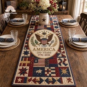 May include: A patriotic table runner with a patchwork design in red, white, and blue. The center features a circular emblem with an eagle and the text "AMERICA 250 YEARS 1776-2026". The runner is set on a wooden table with place settings.