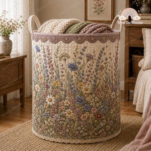 May include: A cylindrical fabric laundry basket featuring a quilted exterior with a colorful wildflower print in shades of blue, yellow, pink, and green. The basket has two top handles, a scalloped trim at the rim, and a lace border at the base.