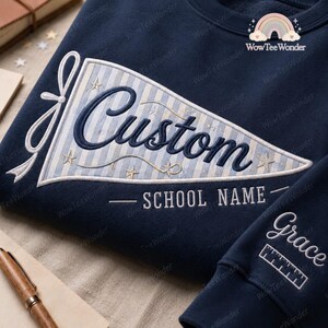 May include: A navy blue sweatshirt featuring a large pennant-shaped patch with light blue and white vertical stripes. The patch displays the word Custom in dark blue script, the text SCHOOL NAME, and small embroidered stars. The sleeve shows the name Grace.