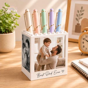 May include: A white square desk organizer made of interlocking building blocks, featuring a photo of a father and child on the front and side. The front panel displays the text Best Dad Ever with a heart. Five pastel-colored pens are stored inside.