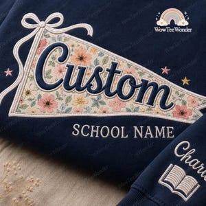 May include: A navy blue sweatshirt featuring an embroidered pennant design with a floral pattern in shades of pink, orange, and green. The word Custom is stitched in dark blue, with the text SCHOOL NAME below. A book icon is on the sleeve.