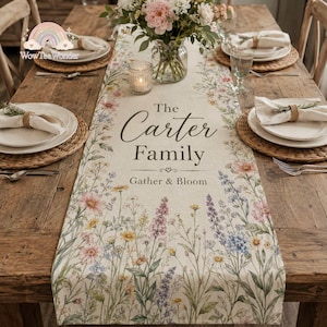 May include: A beige linen table runner featuring a colorful watercolor-style floral print of wildflowers. The text reads The Carter Family and Gather & Bloom. The runner is centered on a wooden table with place settings, plates, and napkins.