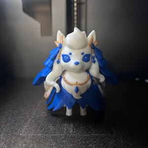 May include: A small white fox-like figurine with blue accents on its ears, eyes, and layered tail. The figure features gold-colored detailing around the ears and a gold chain necklace with a small blue pendant. It stands on four small legs.