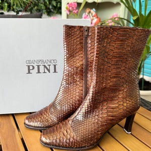 Puede incluir: Un par de botines de cuero marrón texturizado con cierre de cremallera. Los botines tienen punta afilada y tacón pequeño. La marca "GIANFRANCO PINI" está impresa en la caja detrás de los botines.