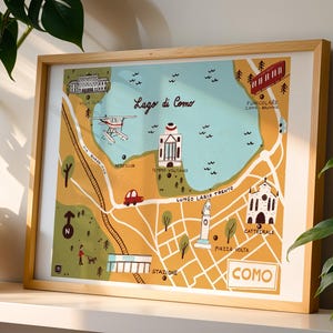 May include: A framed art print featuring a stylized map of Lake Como, Italy. The map is in shades of yellow, blue, and green, with illustrations of landmarks, a seaplane, and a funicular. The word "COMO" is prominently displayed.