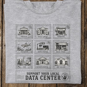 May include: A heather gray t-shirt featuring a black graphic print of nine postage stamp illustrations depicting a theatre, public library, bookstore, aquarium, radio station, music store, zoo, museum, and art gallery, with the text SUPPORT YOUR LOCAL DATA CENTER.