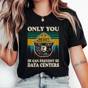 May include: A black short-sleeved t-shirt featuring a graphic of a bear wearing a yellow hat with the word RESIST. The text reads ONLY YOU CAN PREVENT DATA CENTERS above and below the bear, set against a background of colorful horizontal stripes.