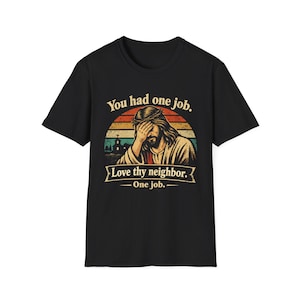 Op de afbeelding: Zwart T-shirt met een afbeelding van Jezus die zijn ogen bedekt, tegen een zonsondergang. De tekst luidt: "You had one job. Love thy neighbor. One job."