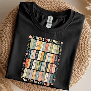 May include: Black t-shirt with the text "FUND LIBRARIES NOT DATA CENTERS" and a colorful graphic of stacked books. The books are decorated with floral and leaf designs.
