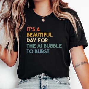 Op de afbeelding: Een zwart T-shirt met korte mouwen en de tekst IT'S A BEAUTIFUL DAY FOR THE AI BUBBLE TO BURST in gestapelde regels. De woorden zijn gekleurd in oranje, geel, crème en lichtblauw, wat zorgt voor een retro-geïnspireerd grafisch ontwerp.