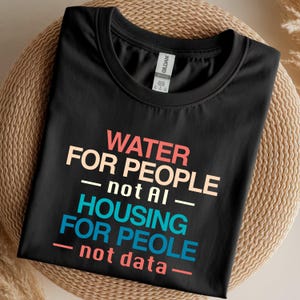 Puede incluir: Una camiseta negra de cuello redondo con texto multicolor que dice: WATER FOR PEOPLE not AI, HOUSING FOR PEOPLE not data. La camiseta tiene una etiqueta de la marca Gildan en el interior del cuello y está hecha de tela negra lisa con un diseño.