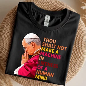 May include: Black t-shirt featuring an image of a religious figure in profile, wearing a white hat and glasses, with hands clasped in prayer. Text on the shirt reads "THOU SHALT NOT MAKE A MACHINE IN LIKENESS OF A HUMAN MIND."