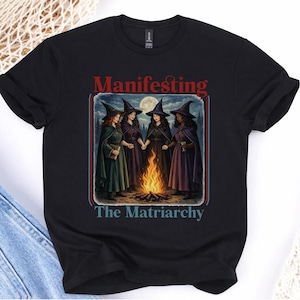 Puede incluir: Camiseta negra con las palabras "Manifesting The Matriarchy" en rojo y azul. El diseño muestra a cuatro mujeres con sombreros y capas de brujas alrededor de una hoguera bajo la luna llena.