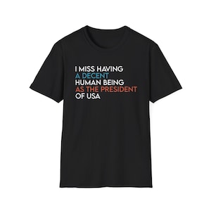 Puede incluir: Una camiseta negra de manga corta con texto en blanco, azul claro y naranja que dice: I MISS HAVING A DECENT HUMAN BEING AS THE PRESIDENT OF USA. La camiseta tiene cuello redondo y un corte estándar, mostrando el texto claramente al frente.