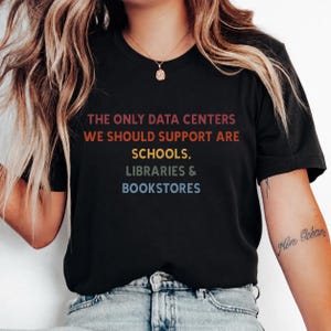 Puede incluir: Camiseta negra con el texto "THE ONLY DATA CENTERS WE SHOULD SUPPORT ARE SCHOOLS, LIBRARIES & BOOKSTORES" en un degradado de colores. La camiseta es de cuello redondo.