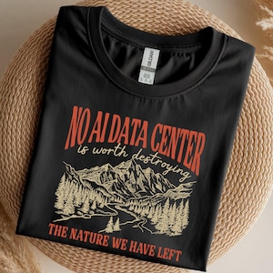 Puede incluir: Una camiseta negra de cuello redondo con un estampado gráfico de una cordillera y un paisaje forestal. El texto dice NO AI DATA CENTER is worth destroying THE NATURE WE HAVE LEFT en colores naranja y crema, con una etiqueta de marca Gildan.